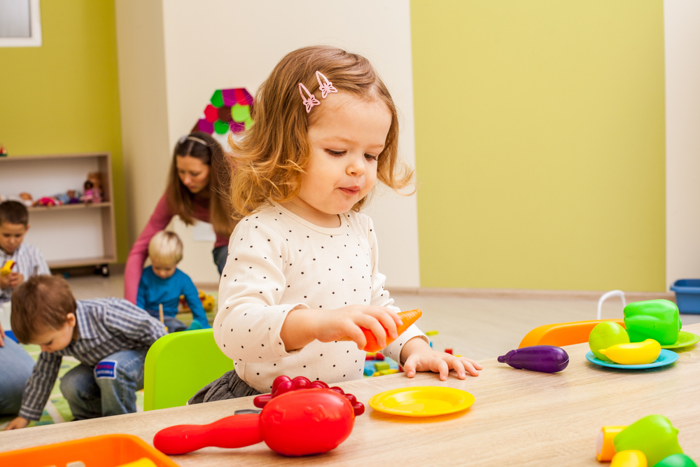 Petite enfance autisme Petite fille blonde autiste qui joue à la dînette avec une salle de jeux avec une éducatrice et d'autres enfants en arrière plan