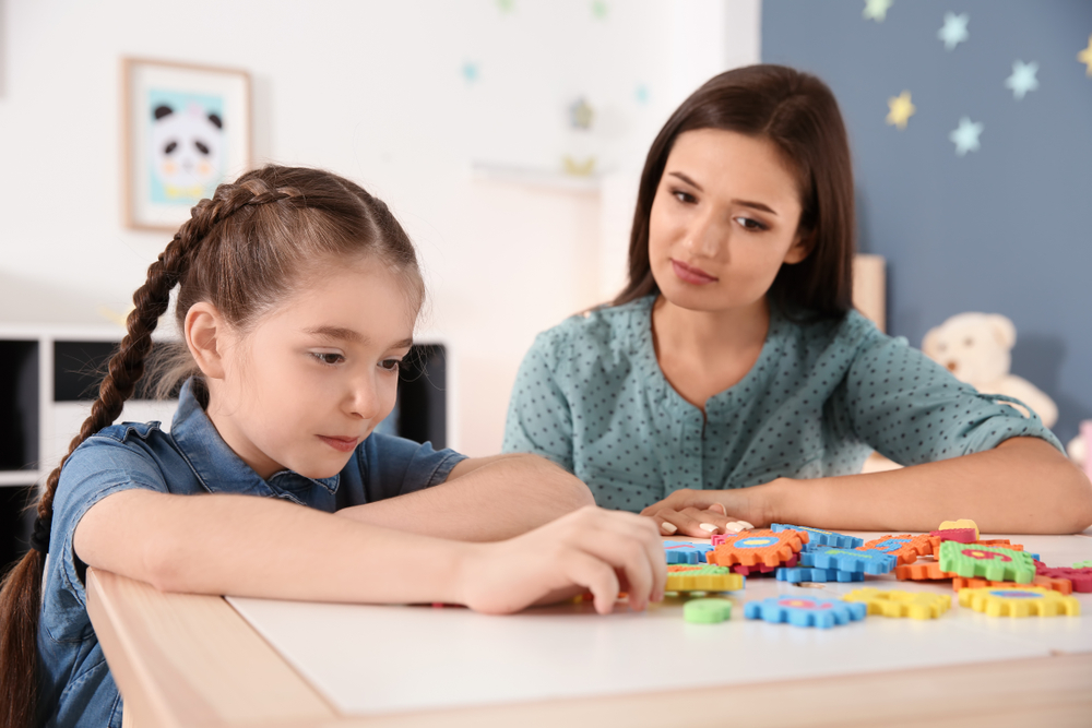 Éducatrice et fillette autiste cote à cote devant des pièces de jeu