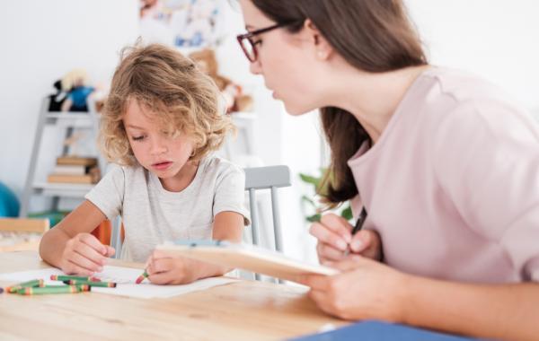 Jeune enfant blond autiste placé à coté d'une educatrice et qui tient un crayon de couleur dans sa main